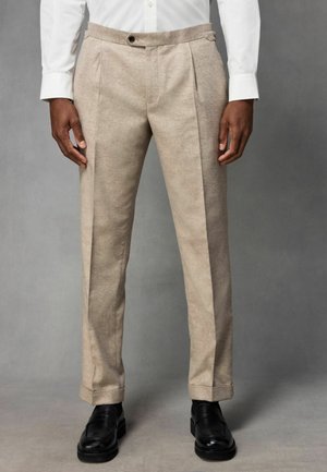 Beige maßgeschneiderte Hose mit texturierter Oberfläche, ausgestattet mit Falten, Seitentaschen und einem umgeschlagenen Saum, kombiniert mit schwarzen Slippern.