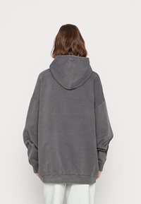 Osoba z włosami brązowymi do ramion, ubrana w luźną, wyblakłą szaro-skalną bluzę oraz białe spodnie, stojąca plecami na jednolitym tle.