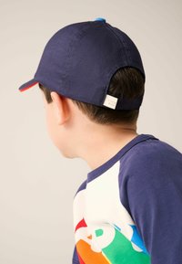 Jūras spēku beisbola cepure ar sarkanu vizu un zilu pogu augšpusē, valkāta atpakaļgaitā. Bērnu krekls ar krāsainu ģeometrisku rakstu baltā un zilā krāsā.