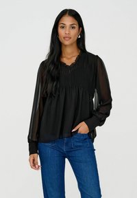 Femme aux longs cheveux noirs portant un chemisier noir transparent à manches longues avec un corsage plissé et un jean bleu, une main dans la poche, debout devant un fond blanc.