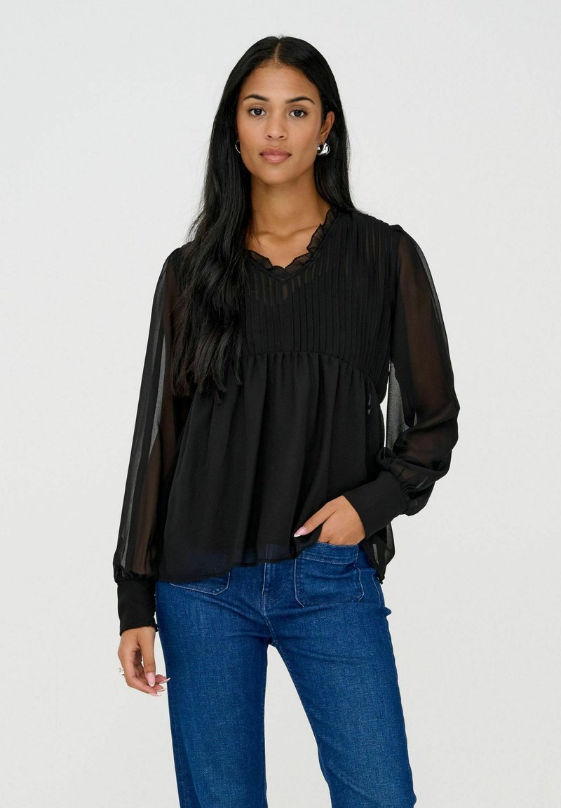 Femme aux longs cheveux noirs portant un chemisier noir transparent à manches longues avec un corsage plissé et un jean bleu, une main dans la poche, debout devant un fond blanc.