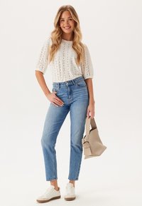 Top bianco in pizzo a maniche corte, jeans blu a vita alta, borsa beige e sneakers bianche. Outfit casual con tagli e texture classiche.