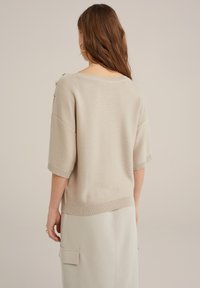 Beige kortärmad stickad tröja med ribbad textur och vid halsringning, med dekorativa knappar på axeln. Enkel och avslappnad passform.