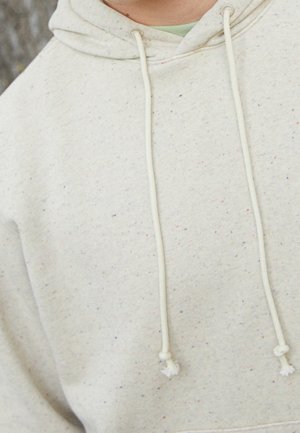 Zbliżenie osoby noszącej beżową, nakrapianą bluzę z kapturem ze sznurkami, pokazujące szyję i górną część klatki piersiowej.