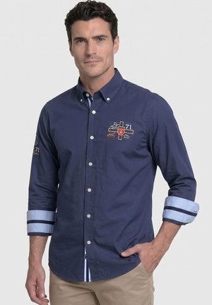 Hombre vistiendo una camisa azul marino con botones, puños azul claro doblados y parches bordados, combinada con pantalones beige.