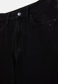 Gros plan d’un jean noir avec des détails subtils pailletés, des poches avant, des passants de ceinture et un bouton argenté marqué « Pull&Bear Since 1991 ».