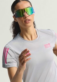 Camiseta deportiva gris con acentos en rosa, que presenta un logo. Gafas de sol grandes y reflectantes con un tinte degradado. Cabello rizado visible.