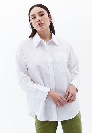 Camicia bianca con bottoni, colletto, maniche lunghe e una tasca sul petto; realizzata in un tessuto liscio; indossata con pantaloni verdi.