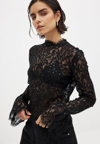 Schwarze Spitzentbluse mit langen, ausgestellten Ärmeln, gewelltem Ausschnitt und verstreuten Pailletten. Der durchsichtige Stoff lässt die darunterliegenden Schichten durchscheinen.