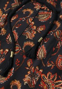 Zwarte stof met een ingewikkeld paisleypatroon in rode, gouden en oranje tinten. Heeft franjes bij de halslijn, wat textuur en detail toevoegt.