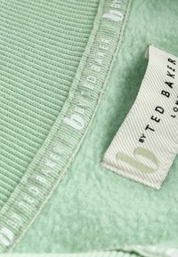 Světle zelená tkanina s texturovaným žebrováním a měkkou, huňatou částí. Obsahuje štítek se slovem "Ted Baker" ve světlejším písmu.