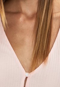 Närbild av en kvinnas bröst med rakt blont hår och en ljusrosa ribbad topp som är knäppt med en pärlknapp.