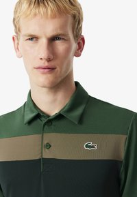 En blond man som bär en randig polo-skorta i grönt, brunt och mörkgrön med en krokodil-logotyp på bröstet, som tittar åt sidan mot en vit bakgrund.