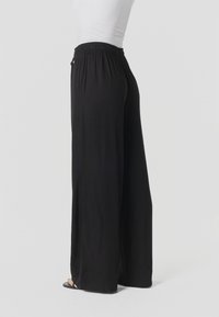 Mujer con pantalones negros de tiro alto y pierna ancha, y top blanco ajustado, de pie descalza frente a un fondo neutro.