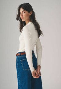 Geribbelde witte top met lange mouwen, gecombineerd met high-waisted donkerblauwe denim jeans en een bruine riem, die een getailleerde silhoutte en soepele textuur laat zien.