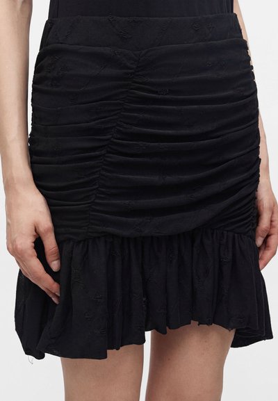 Personne portant une mini-jupe noire froncée avec un ourlet à volants, les mains le long du corps, montrée de la taille à mi-cuisse sur un fond uni.