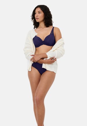 Granatowy biustonosz i pasujące bikini wykonane z gładkiego materiału, ozdobione delikatną koronką oraz biały, teksturowany kardigan noszony opuszczony na ramionach.