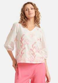Witte blouse met roze bloemenpatroon, V-hals en driekwart pofmouwen, gecombineerd met roze broek. De blouse heeft een lichte stof en een zachte textuur.
