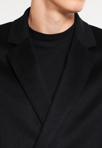 Cappotto nero in misto lana con design a revers classico, caratterizzato da una texture liscia e una forma strutturata, indossato sopra un dolcevita nero.