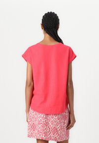 Rotes Baumwollmischgewebe-Oberteil mit kurzen Ärmeln und rundem Ausschnitt, getragen mit einem gemusterten weißen und rosa Rock. Glatte Textur, lässige Passform.