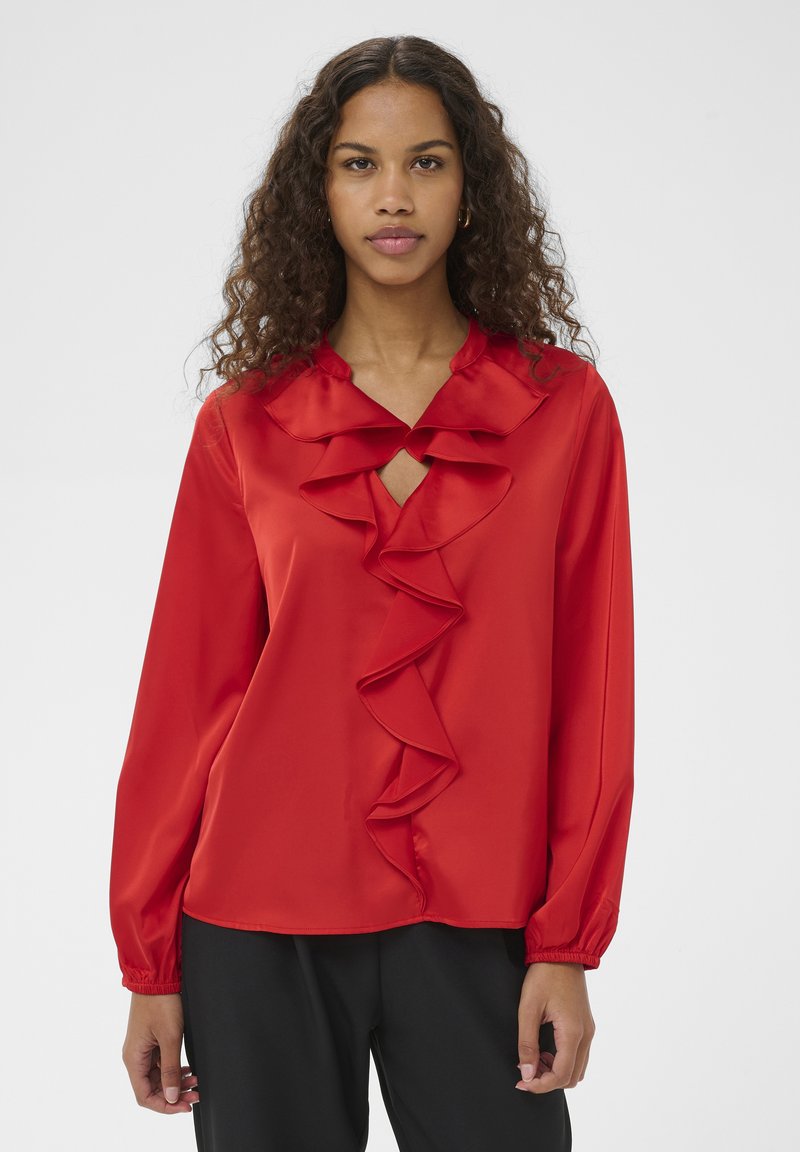 Blouse en satin rouge avec un col en V et un large volant sur le devant. Manches longues avec poignets élastiques. Texture lisse, coupe décontractée.