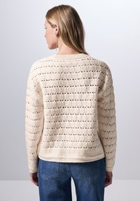 Cremefarbener Strickpullover mit einem strukturierten Muster aus offenen Maschen, lockerer Passform, Rundhalsausschnitt und gerippten Bündchen, kombiniert mit blauen Jeans.