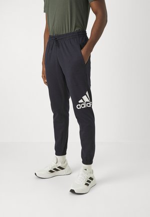 Mann trägt schwarze Adidas Jogginghosen mit großem weißem Logo am linken Oberschenkel und weiße Adidas-Sneaker vor einfarbigem Hintergrund.