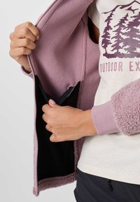 Hands holding open a mauve fleece jacket to show a black inner pocket, worn over a white shirt with purple tree design and text.