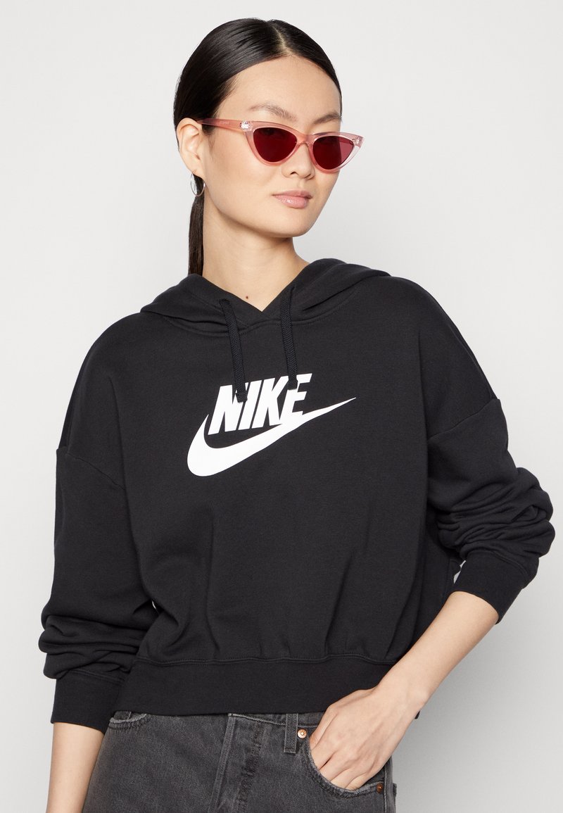 Sudadera corta negra con un gran logo blanco de Nike, con capucha ajustable y puños acanalados. Modelo llevando gafas de sol rosa en forma de gato.