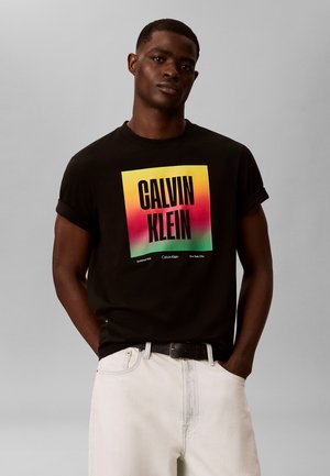 Άνδρας που φοράει μαύρο μπλουζάκι Calvin Klein με πολύχρωμο τετράγωνο λογότυπο και λευκό παντελόνι, στέκεται με τα χέρια στις τσέπες μπροστά σε γκρι φόντο.