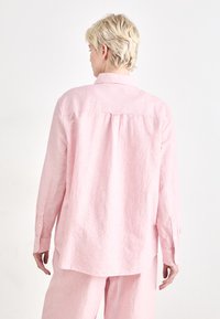 Light pink button-up shirt made of textured fabric, featuring a back pleat, long sleeves, and rounded hem. Visible cuff and button detail.