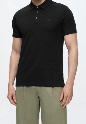 Man draagt een zwart poloshirt met korte mouwen en knopen en lichtgroene korte broek, staat voor een effen achtergrond.