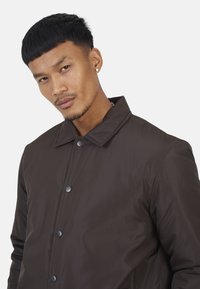 Veste marron à boutons, avec un col, dont la texture du tissu semble lisse et mate. Elle comporte des boutons foncés et un design décontracté.