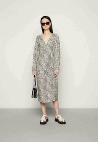 Vestido midi de estampa de leopardo com mangas longas, decote em V, cintura ajustada e saia esvoaçante. Combinado com sandálias brancas e pretas e uma bolsa preta.