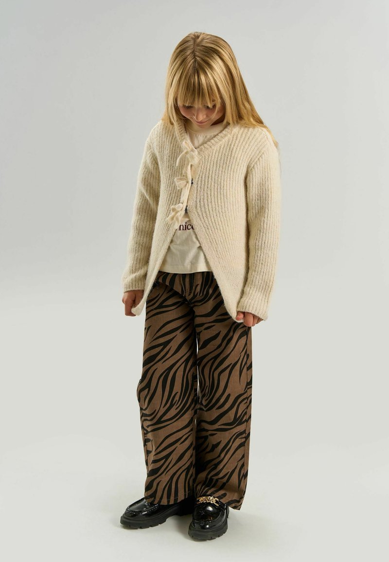 Jeune enfant portant un cardigan en maille crème, un t-shirt graphique beige, un pantalon large marron à motif zèbre et des mocassins noirs avec un détail chaîne dorée, regardant vers le bas.