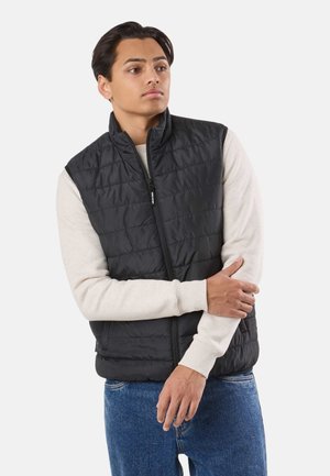 Młody mężczyzna ubrany w czarną pikowaną kamizelkę na kremowej, długorękawowej koszuli i niebieskich dżinsach, z założonymi rękami, patrzący w bok.