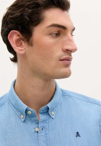 Lichtblauwe denim knoopshirts met een klassieke kraag, met een klein donker logo dicht bij de borst en zichtbare stikdetails.