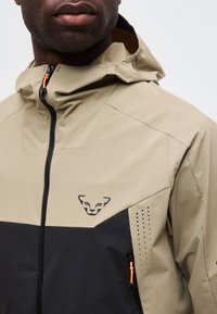 Κοντινό πλάνο ενός ατόμου που φοράει μπεζ και μαύρο μπουφάν με κουκούλα, με μαύρο λογότυπο ζώου στο στήθος και πορτοκαλί τράβηγμα φερμουάρ.