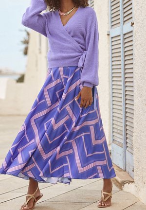 Femme portant un pull en cachemire lavande et une jupe fluide bleue avec un motif géométrique rose, debout près d'un mur blanc et d'une fenêtre à volets.