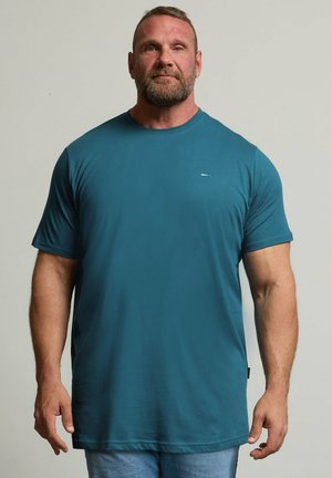 Muskelmand med kort hår og skæg iført en ensfarvet blå kortærmet t-shirt og lyseblå jeans mod en ensfarvet baggrund.