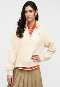Φούτερ με φερμουάρ στο χρώμα της κρέμας και ψηλό γιακά, με μπλε λογότυπο. Φορεμένο πάνω από πορτοκαλί και λευκή ριγέ πόλο, συνδυασμένο με φούστα με πιέτες.