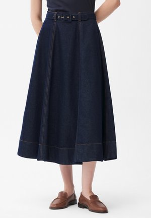 Frau trägt einen dunkelblauen Midilänge-Denimrock mit passendem Gürtel und braunen Leder-Loafers, steht vor einem weißen Hintergrund.