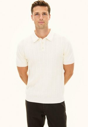 Mężczyzna o jasnobrązowych włosach, ubrany w biały dzianinowy polo z krótkim rękawem i czarne spodnie, stoi z rękami za plecami.