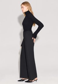 Kobieta ubrana w czarny dopasowany golf, czarne spodnie o wysokim stanie i szerokich nogawkach oraz czarne szpilki na pięcie, stojąca w profilu.