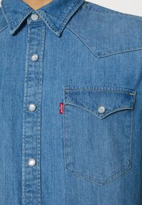 Ljusblå denimskjorta med spetsig krage, silverfärgade tryckknappar och en bröstficka. Anmärkningsvärt Levi's-röd märke på vänster ficka.