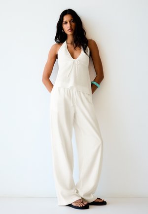 Femme debout contre un mur blanc, portant un haut blanc sans manches à col halter, un pantalon blanc ample, des sandales noires et un bracelet turquoise.