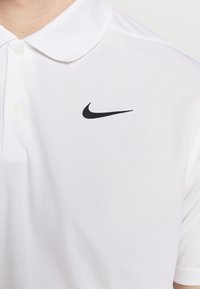 Bílá polo košile vyrobená z lehkého materiálu s klasickým límcem. Na levé straně hrudi se nachází černé logo Nike. Viditelné jsou dva knoflíky.