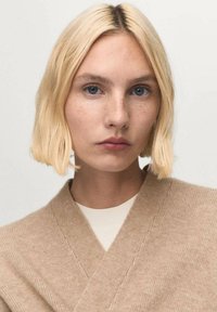 Giovane donna con capelli biondi corti, occhi azzurri, leggeri lentiggini, che indossa un maglione di maglia beige sopra una maglietta bianca, rivolta in avanti con un'espressione neutra.