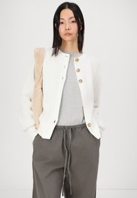 Cardigan en tricot blanc avec trois grands boutons beiges, porté par-dessus un haut gris, associé à un pantalon gris ample. Tissu texturé, coupe décontractée.