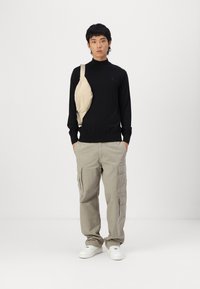 Camicia nera a maniche lunghe, borsa a tracolla beige, pantaloni cargo grigi chiari e sneakers bianche; il modello è in piedi con le mani in tasca.